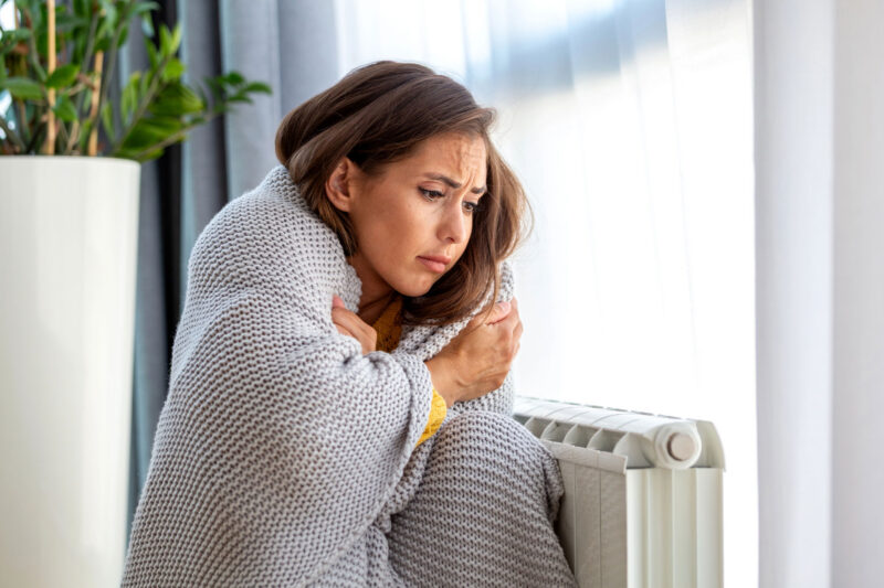 Frierende Frau sitzt mit einer Decke neben einem Heizkörper in einer kalten Wohnung.
