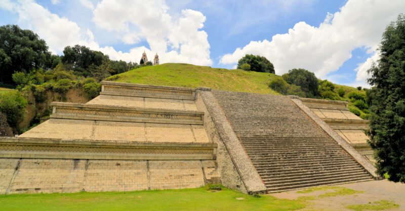 Gro&szlig;e Pyramide von Cholula in Mexiko mit breiter Treppenanlage, gestuften Mauerfl&auml;chen und bewachsenem H&uuml;gel im Hintergrund.