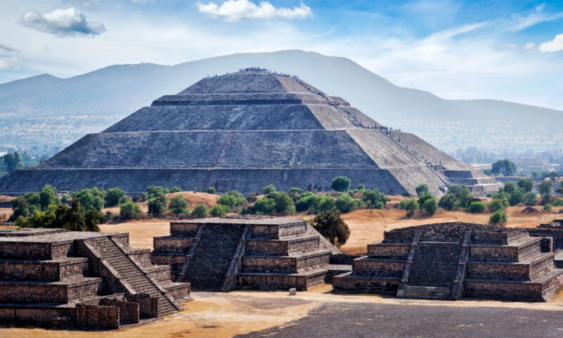 Sonnenpyramide von Teotihuac&aacute;n in Mexiko mit breiten Stufen, vorgelagerten kleineren Tempelbauten und Bergen im Hintergrund.